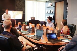 Coworkers sit around a conference table with laptops while a colleague presents at a screen.