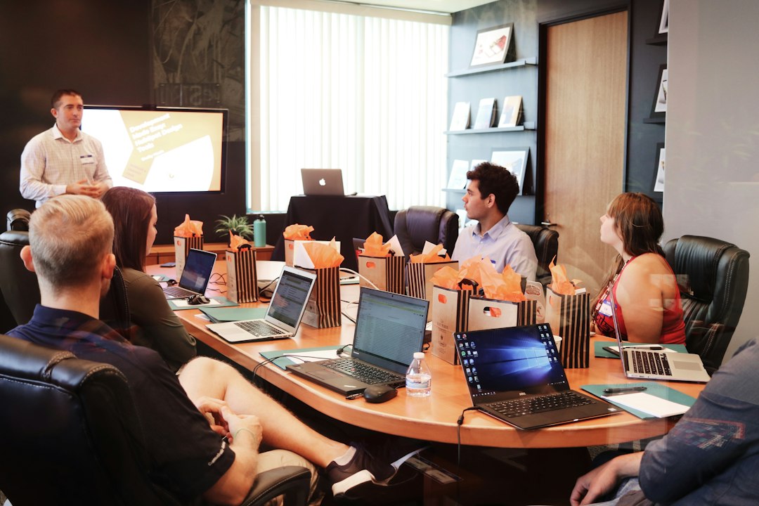 Coworkers sit around a conference table with laptops while a colleague presents at a screen.