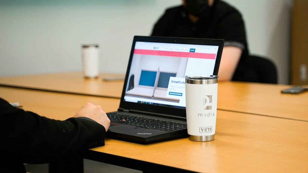 Person typing on a laptop showing a website homepage, with a white tumbler beside it on a conference table.