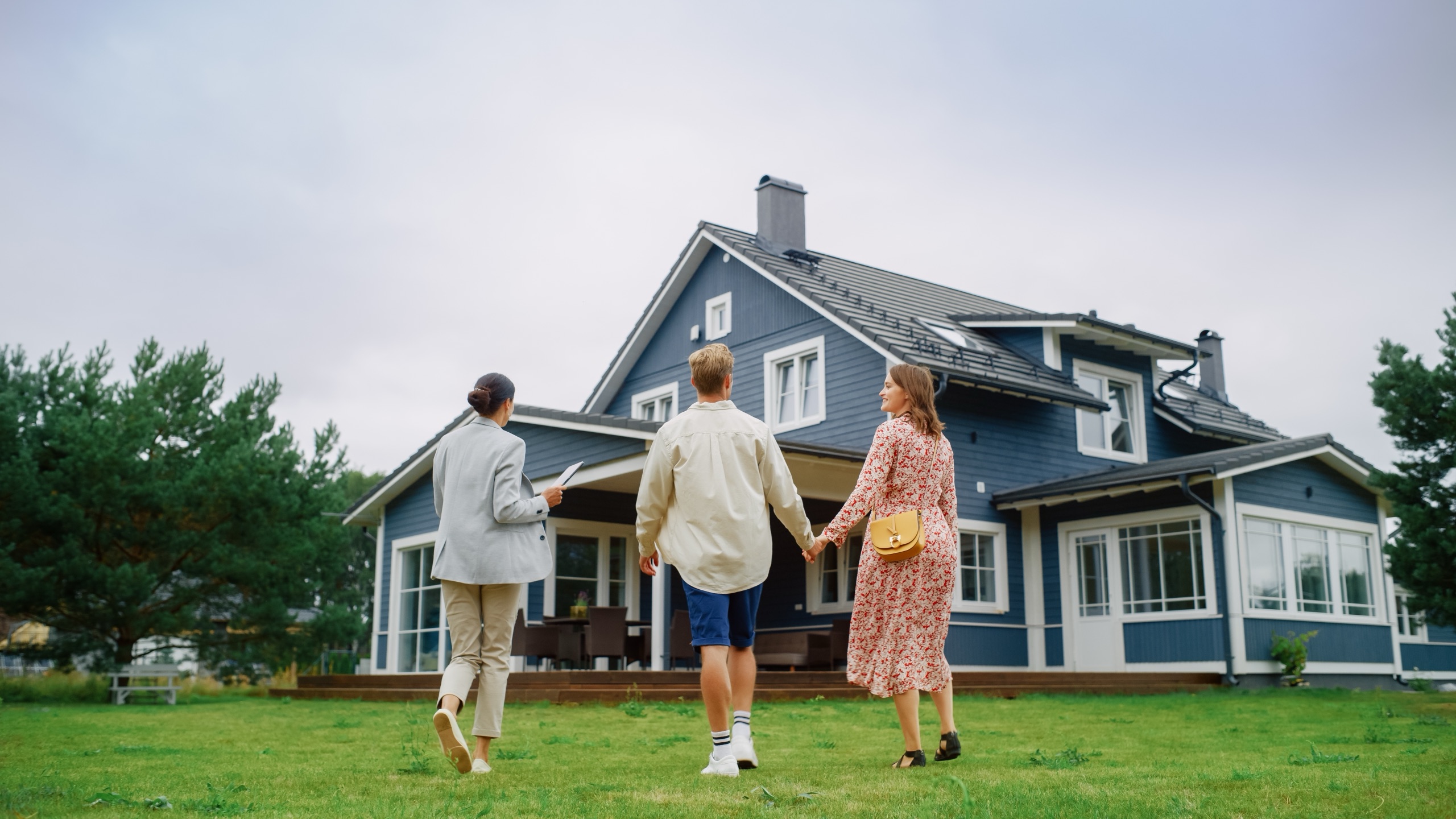 A couple walking hand in hand with a real estate agent toward a blue two-story house.