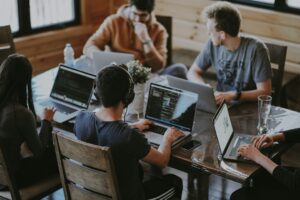 Five people work on laptops around a wooden table, one wearing headphones and writing code.