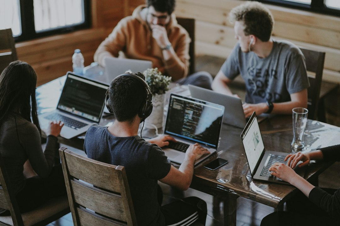 Five people work on laptops around a wooden table, one wearing headphones and writing code.