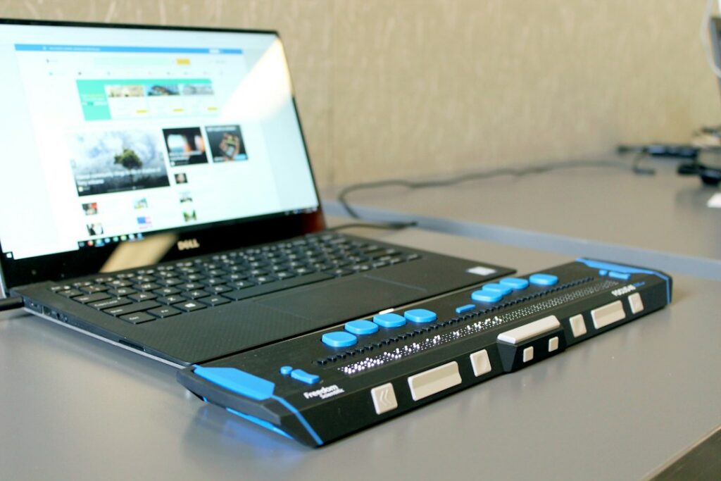 A refreshable braille display sits in front of an open laptop showing a website.